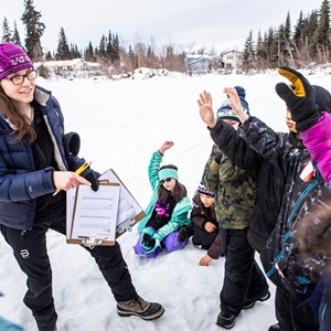 Photo of a woman in cold weather gear holding a clipboard and talking to students outdoors on the snow covered ground.