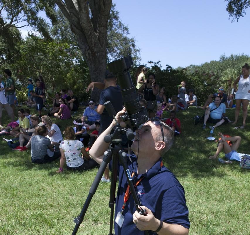 A man in a blue shirt crouches on grass. He is holding a large camera up to his eye and is pointing it at the sky. He has a large smile on his face. Behind him, many people are gathered in the shade of a tree.