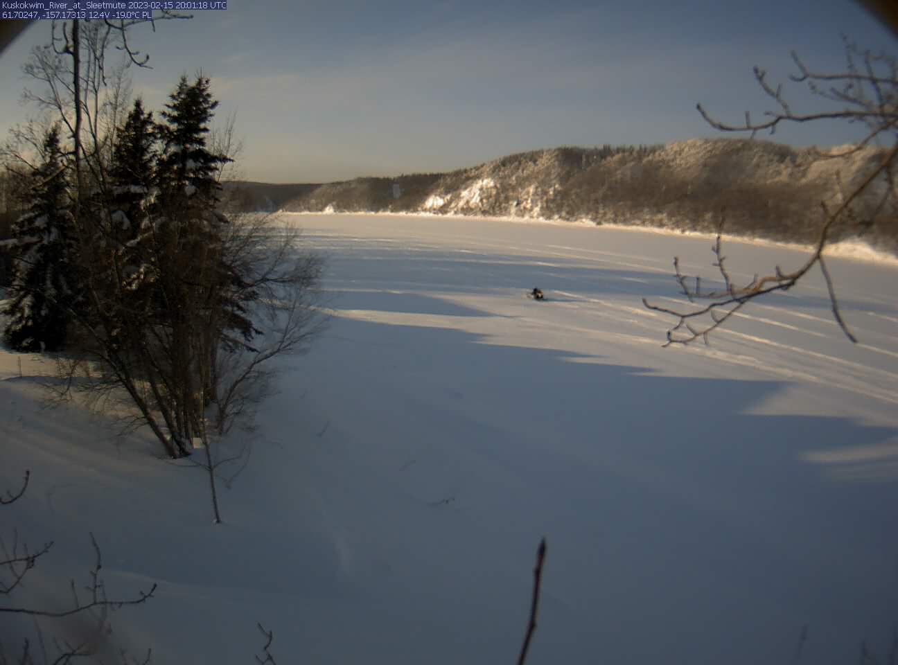 a lone snowmobile, tiny in the big landscape, travels away from us on an ice and snow-covered river. Low sunlight casts long shadows out onto the snow of the pine trees lining the riverbank. The sky is pale blue. The tree-covered hills on the far side of the river have fresh snow in their branches.