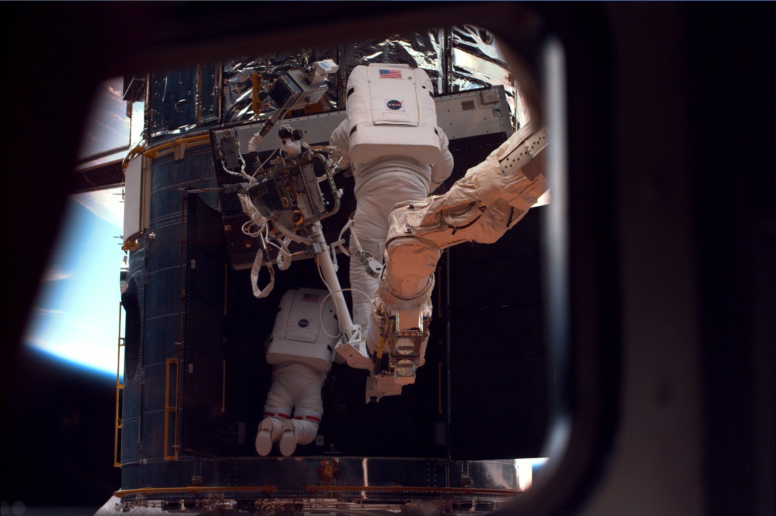One astronaut perches on the robotic arm and another is only partially visible inside a dark open door on the Hubble Space Telescope. Both astronauts are seen from behind, with Hubble looming before them.
