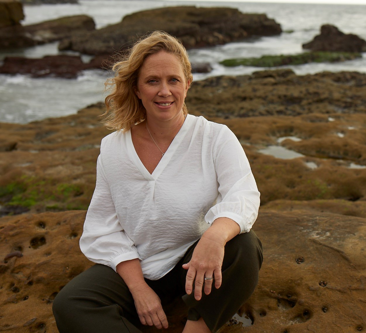 Centered in the image is a woman wearing a white linen shirt and dark pants. She is sitting on a rock, her knees tucked up as if she is in a crouching position. She has blonde hair which is blowing slightly with the breeze to the left of the image. The rock she is sitting on is brown colored and has small craters in it. The rocky landscape extends behind her where it then reaches the ocean, which is a light cray color. There are rocks extending from the ocean as well.