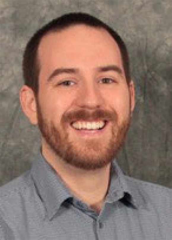 Head shot of Gregory Mace, male, with a big smile and a reddish-brown beard, black hair, gray shirt.