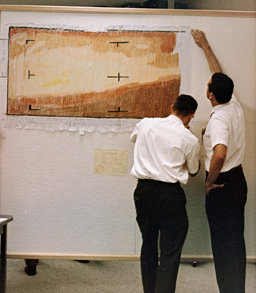 Two men color in data on strips of paper mounted to a wall.