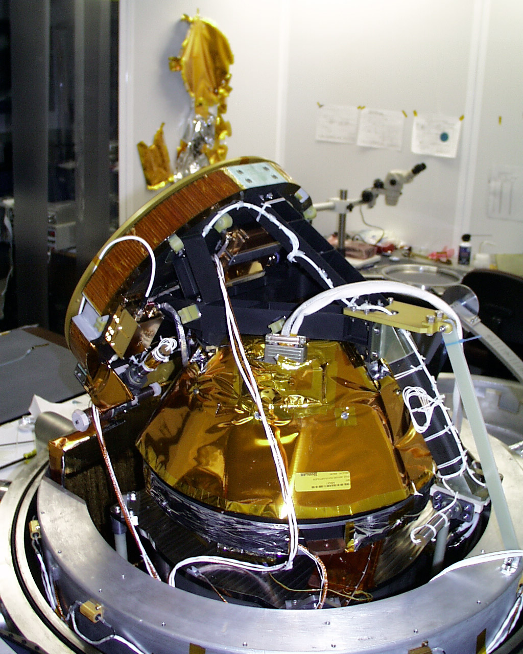 A black and silver cone-shaped device, wrapped in gold foil around its center, sits atop a dull gray ring on a table in a workshop or lab. Instead of a point, its top is short, squat, cylinder, and veers off-center, toward the upper left of the image at a 120-degree ange. The cylinder is also gold around its top edge, which is reeded like the edge of a coin, and the entire device has white wires plugged into it at various locations.