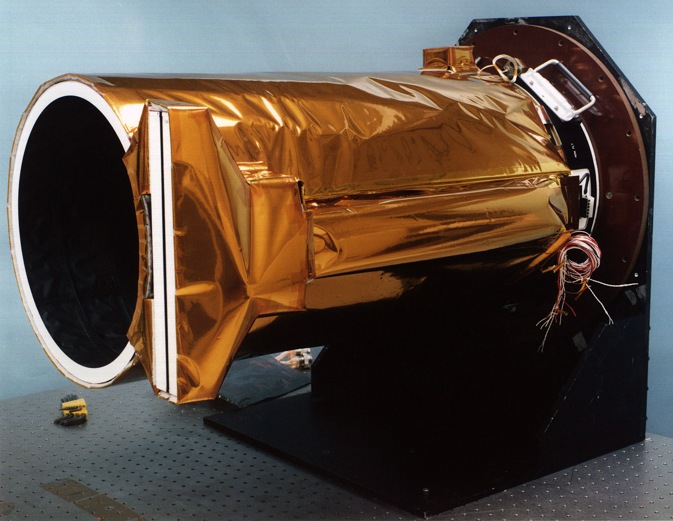 A long, metallic, barrel-shaped instrument, oriented horizontally, points toward the left side of the frame, on a slight diagonal toward the viewer, showing a pitch-black opening that looks like the hole of a deep well. Its sides are covered in what looks like thin, gold foil, it has a bundle of wires protruding from its base, and it's attached to a circular brown metal base at the right rear of the image.