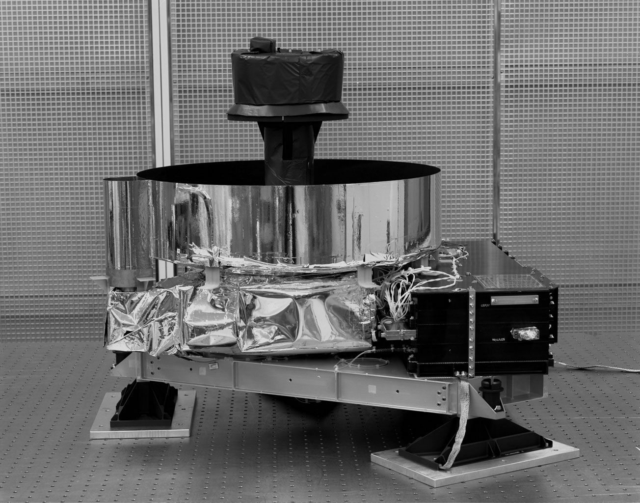 A black-and-white photo of a science instrument, consisting of a metal platform elevated atop three retangular feet. Above the platform rises a squat silver cylinder with an open top and mirrored outside. Sicking up from inside the cylinder is a black, mushroom-shaped element. The base of the cylinder is wrapped in silver foil, and a black box also rests on the platform, to the right.