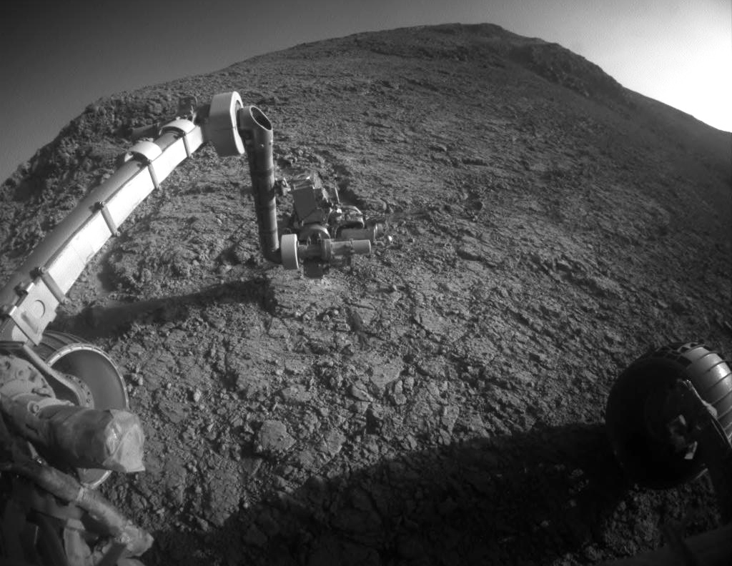 A robotic arm extends over rough terrain on Mars.