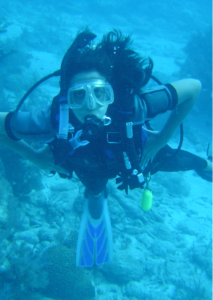 A woman is seen underwater, giving the entire picture a blue hue. She is wearing a scuba mask and has a respirator in her mouth, connected to a tank on her back with several tubes. She is wearing flippers and a wet suit. Her dark hair is floating all around her head as she looks up at the camera. Behind her is a coral reef and sand at the bottom of the ocean can be seen to the right of the image too.