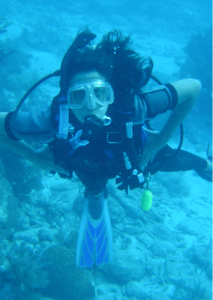 A woman is seen underwater, giving the entire picture a blue hue. She is wearing a scuba mask and has a respirator in her mouth, connected to a tank on her back with several tubes. She is wearing flippers and a wet suit. Her dark hair is floating all around her head as she looks up at the camera. Behind her is a coral reef and sand at the bottom of the ocean can be seen to the right of the image too.