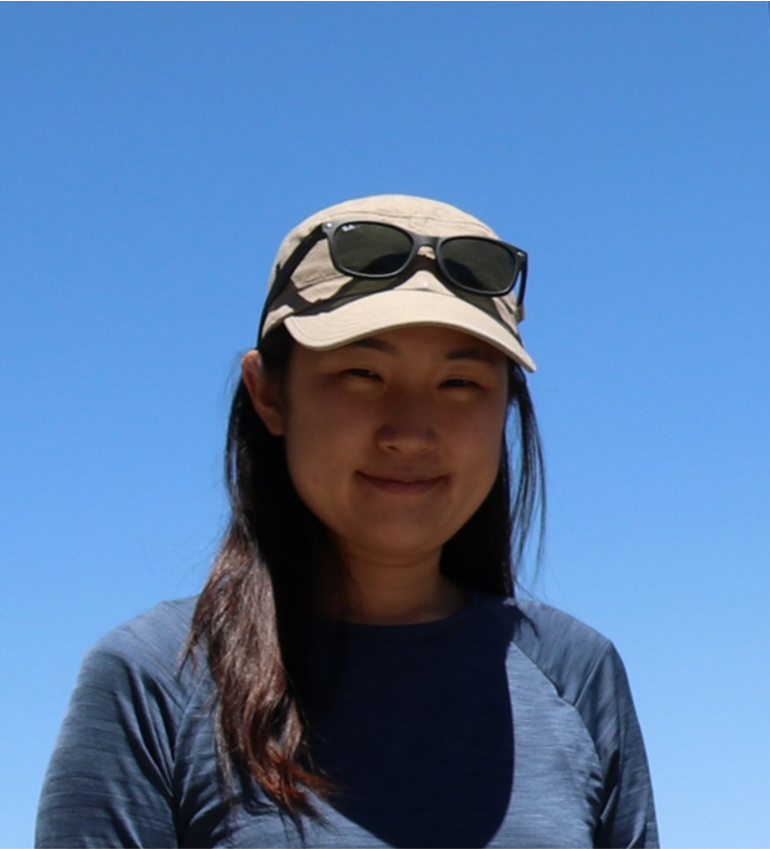 Portrait photo of a young woman outside against a bright blue sky; she is wearing a cap with her sunglasses sitting on top of it.