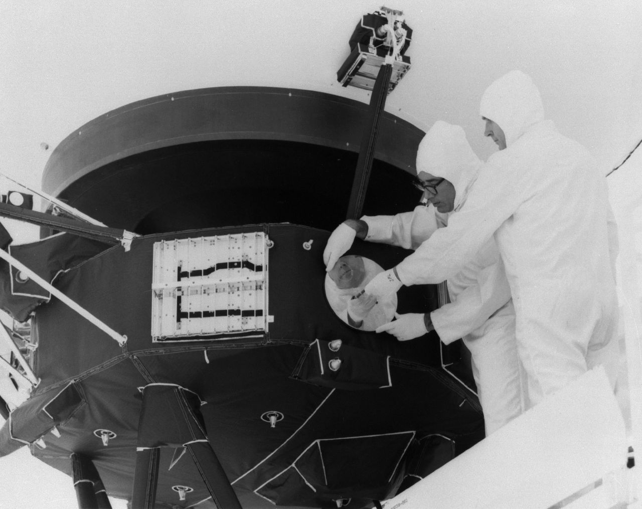 Two men in white cleanroom suites attach the golden record to the side of the Voyager spacecraft.