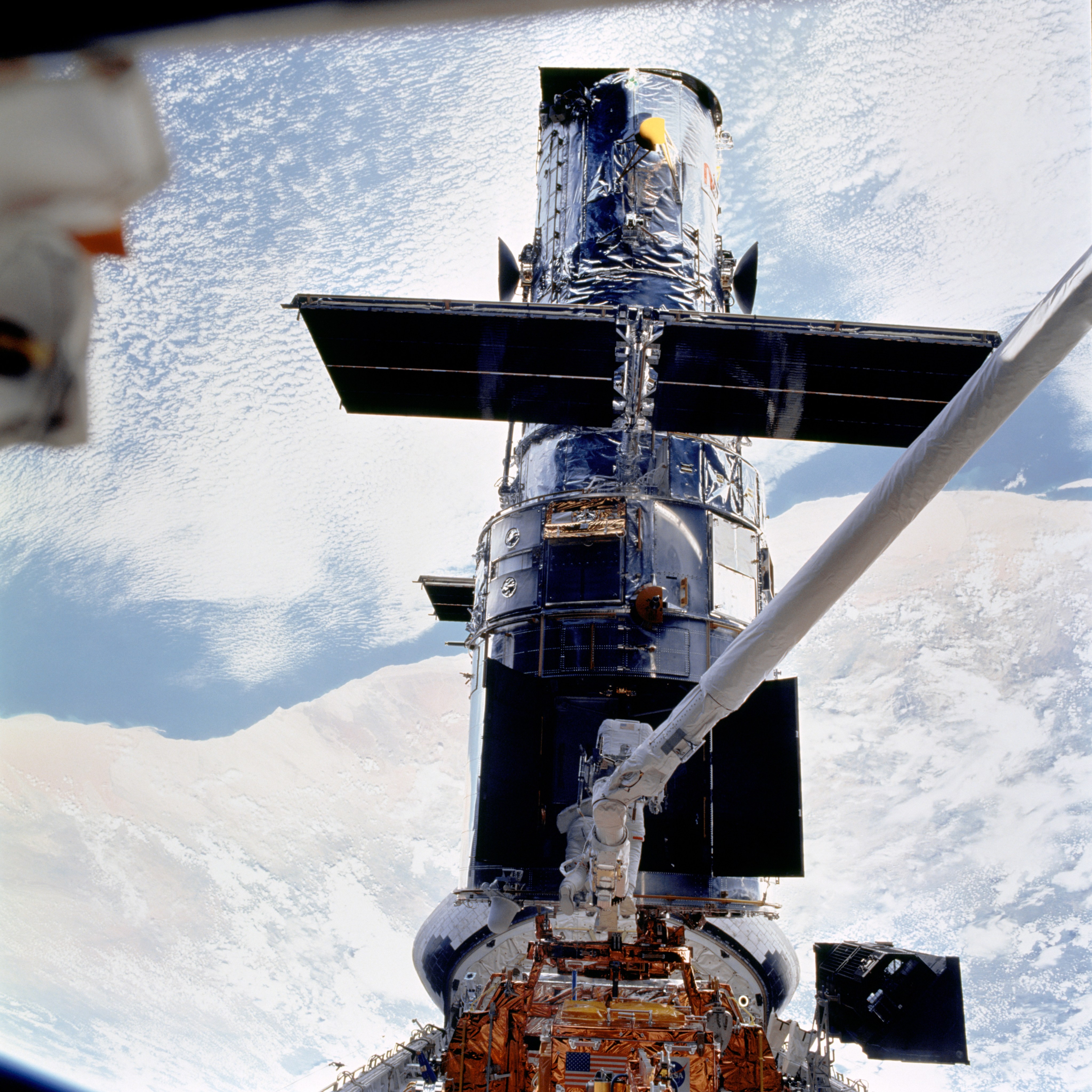 Hubble is framed by a blue and cloudy Earth as astronauts work on the telescope, one perched on the shuttle's robotic arm.