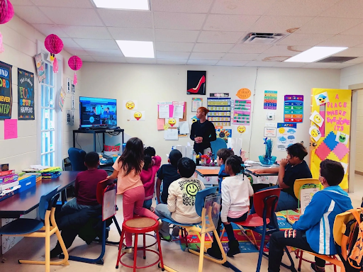 A classroom photo with seated children focused on their teacher standing at the front of the room. The walls are filled with colorful projects, artwork and decorations.