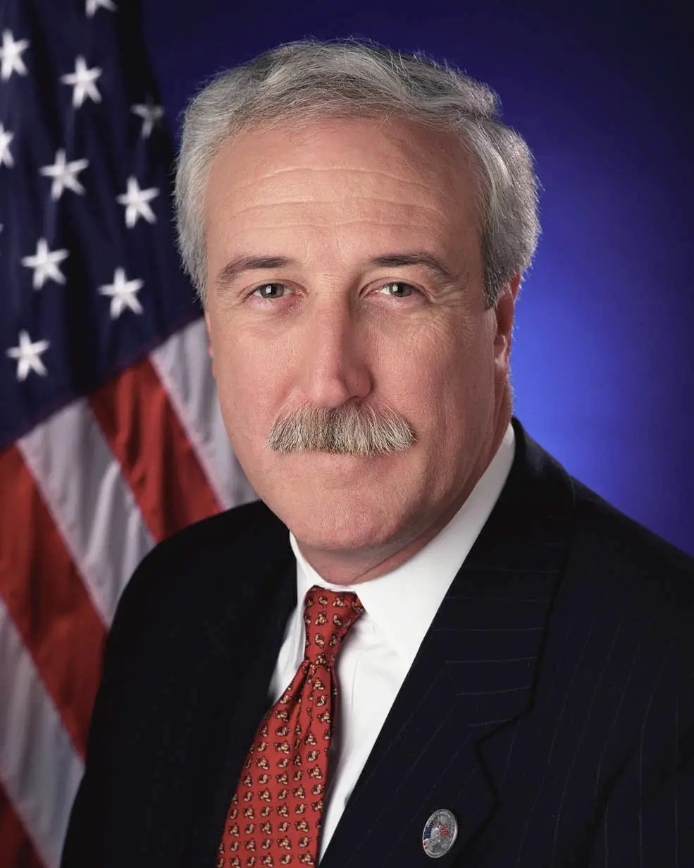 Headshot of a man with white hair and mustache wearing a navy blue jacket, white shirt and red tie.