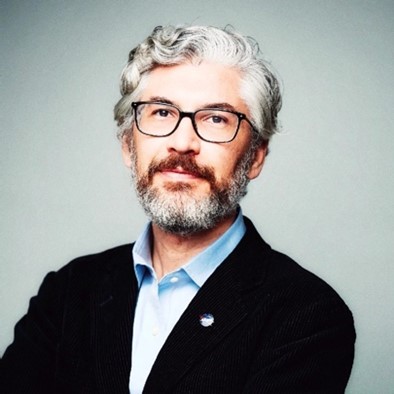 Mid-body and head shot photograph of Dr. Omid Noroozian, wearing a black jacket and light blue buttoned down shirt and a NASA pin on his left, male with gray/white/black hair and full beard, wearing glasses with black frames.