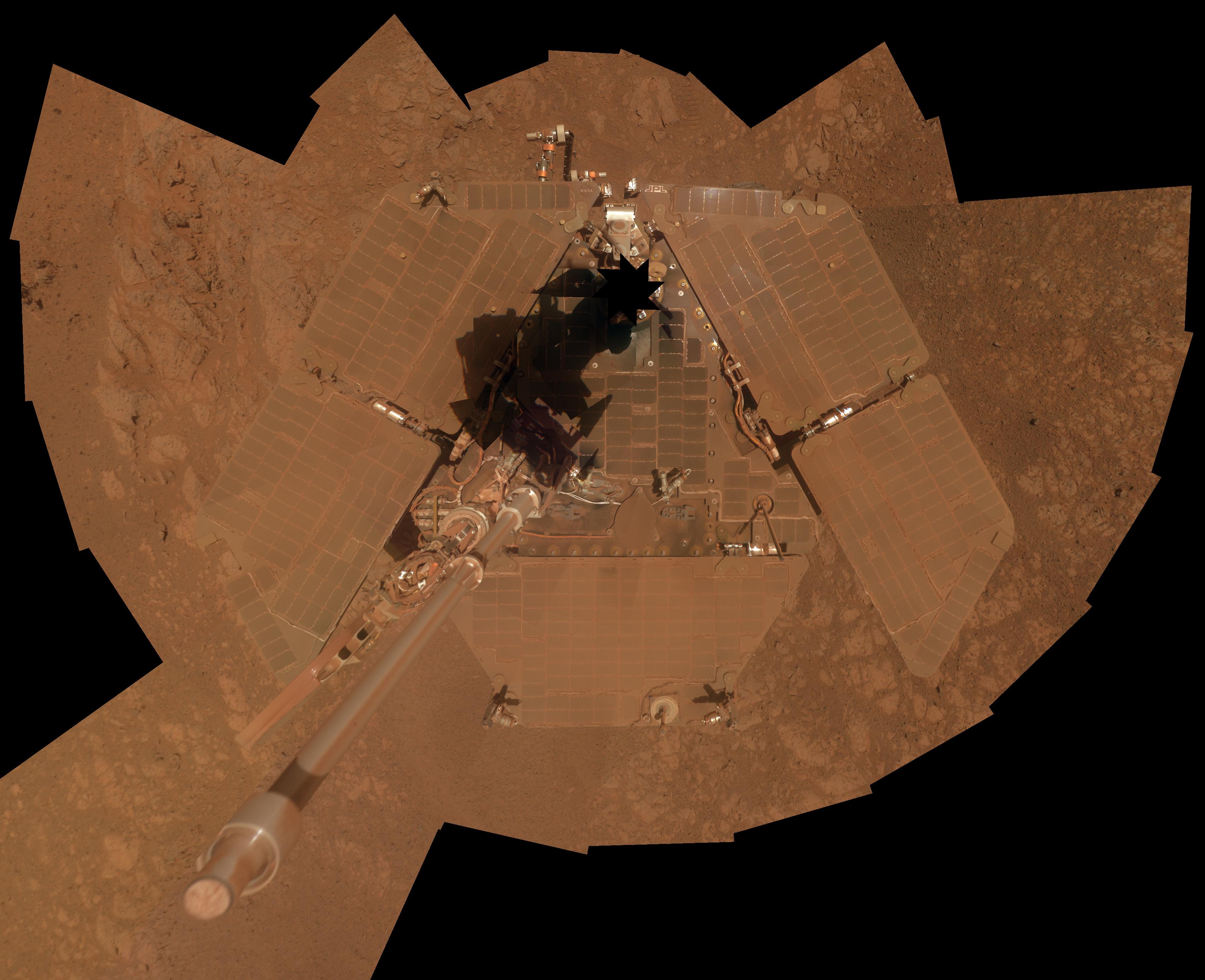 An overhead view of a Mars rover, of which you can only see its triangular top deck, at the center of the image pointing toward the top of the frame, and the deck is surrounded by five trapezoidal solar panels – two each extending from the top two sides of the triangle, and the fifth one extending from the triangle's base. But the entire thing is so covered in cinnamon-colored dust, it's almost impossible to distinguish from the surrounding terrain. A silvery mast, also dust-covered, extends upward from the deck toward the viewer.