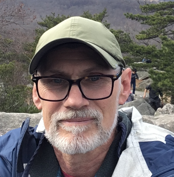 Portrait photo of man in a cap wearing glasses in and outdoor setting.