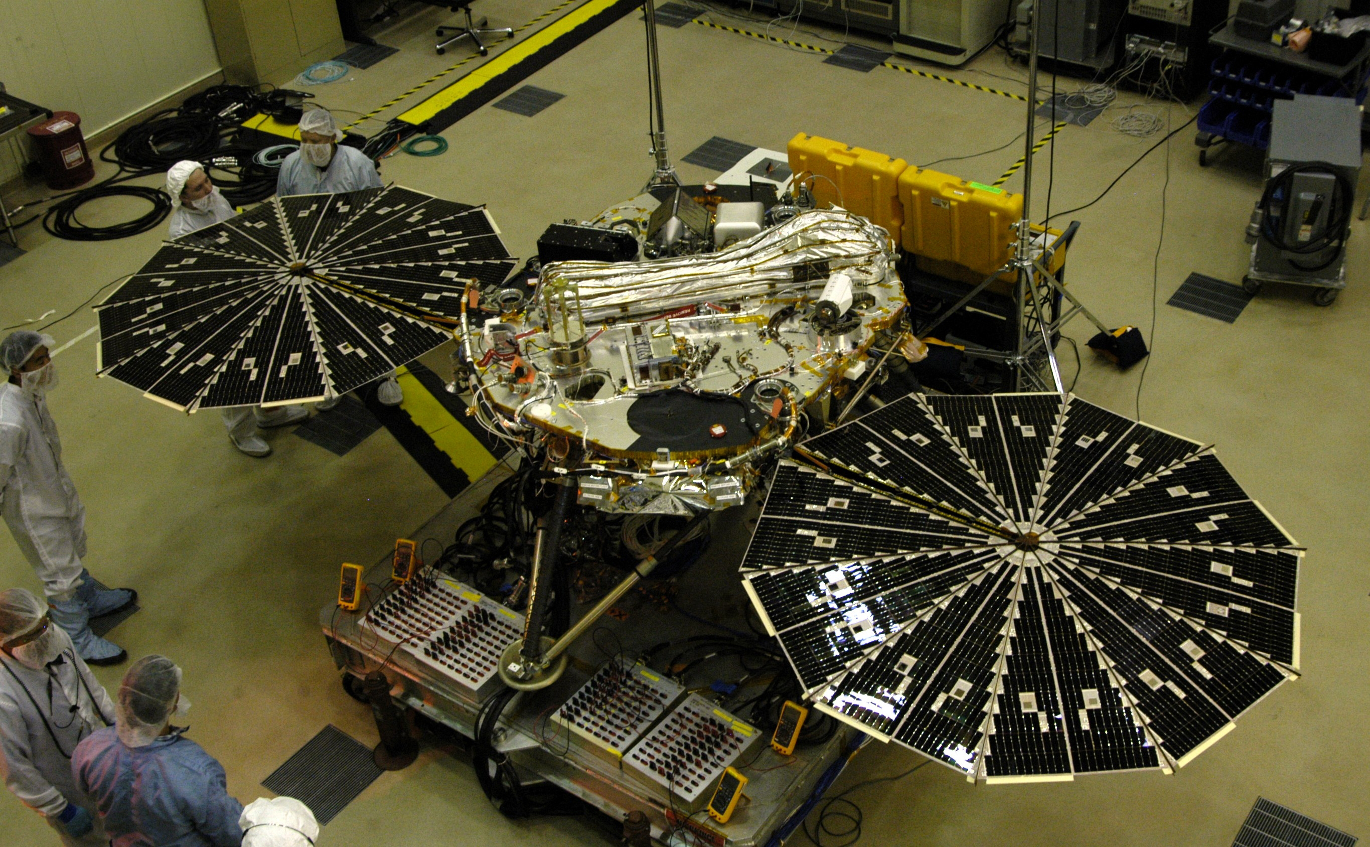 A four-legged spacecraft is sitting in a cleanroom with its solar panels wide open.