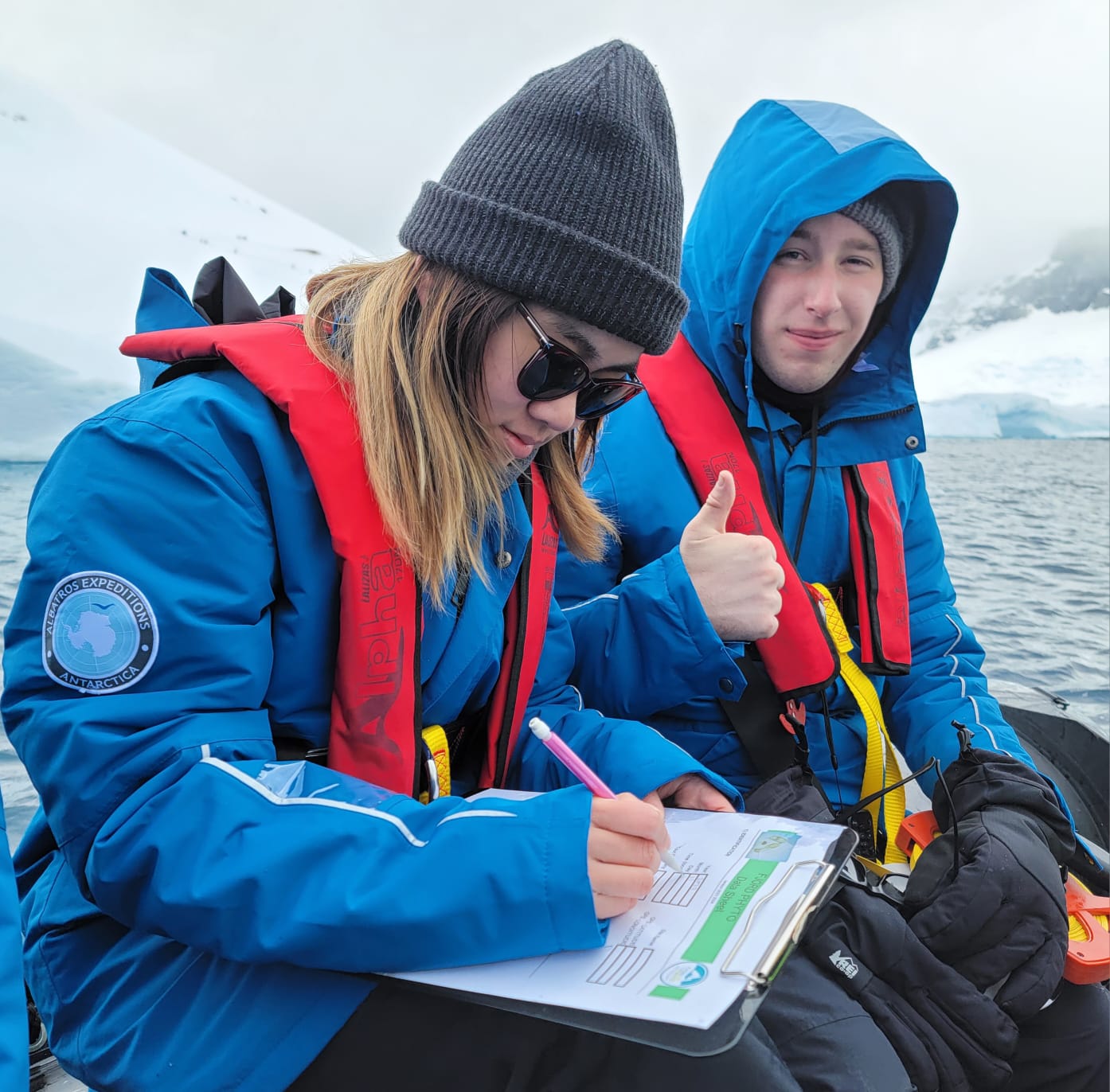 Photo of a young man and woman in blue winter jackets, red floatation vets and warm hats sitting on a boat in the Antarctic. The woman is writing