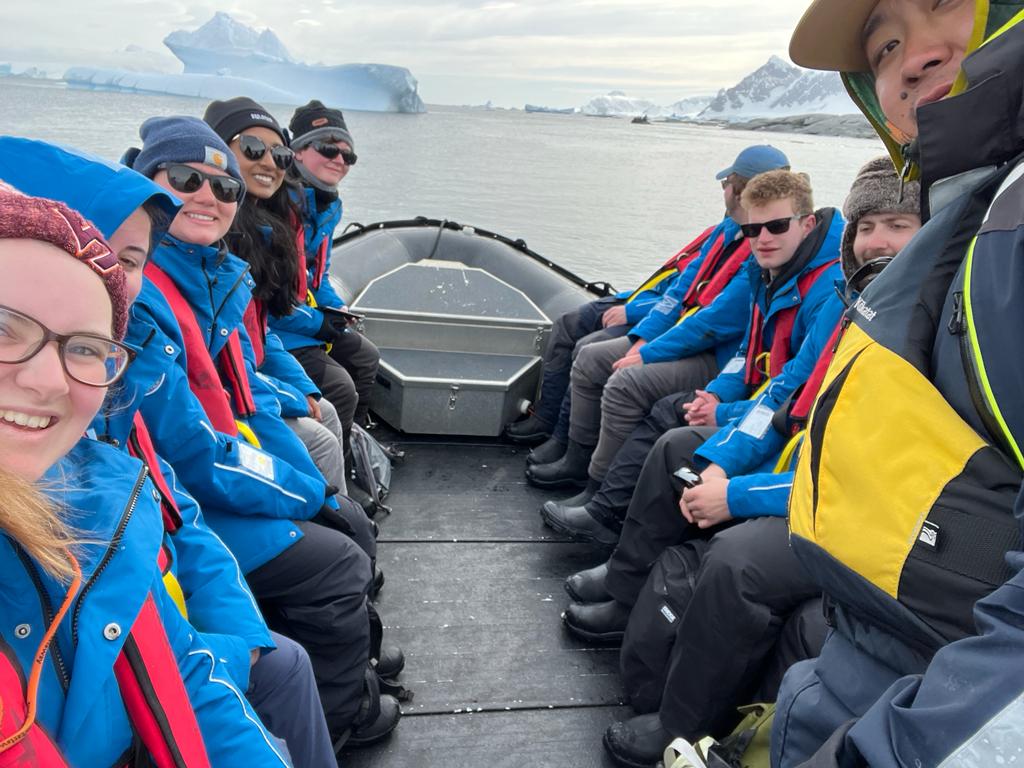 Photo of a group of 10 people, most wearing blue winter jackets and red floatation vests, sitting on a boat in the Antarctic.
