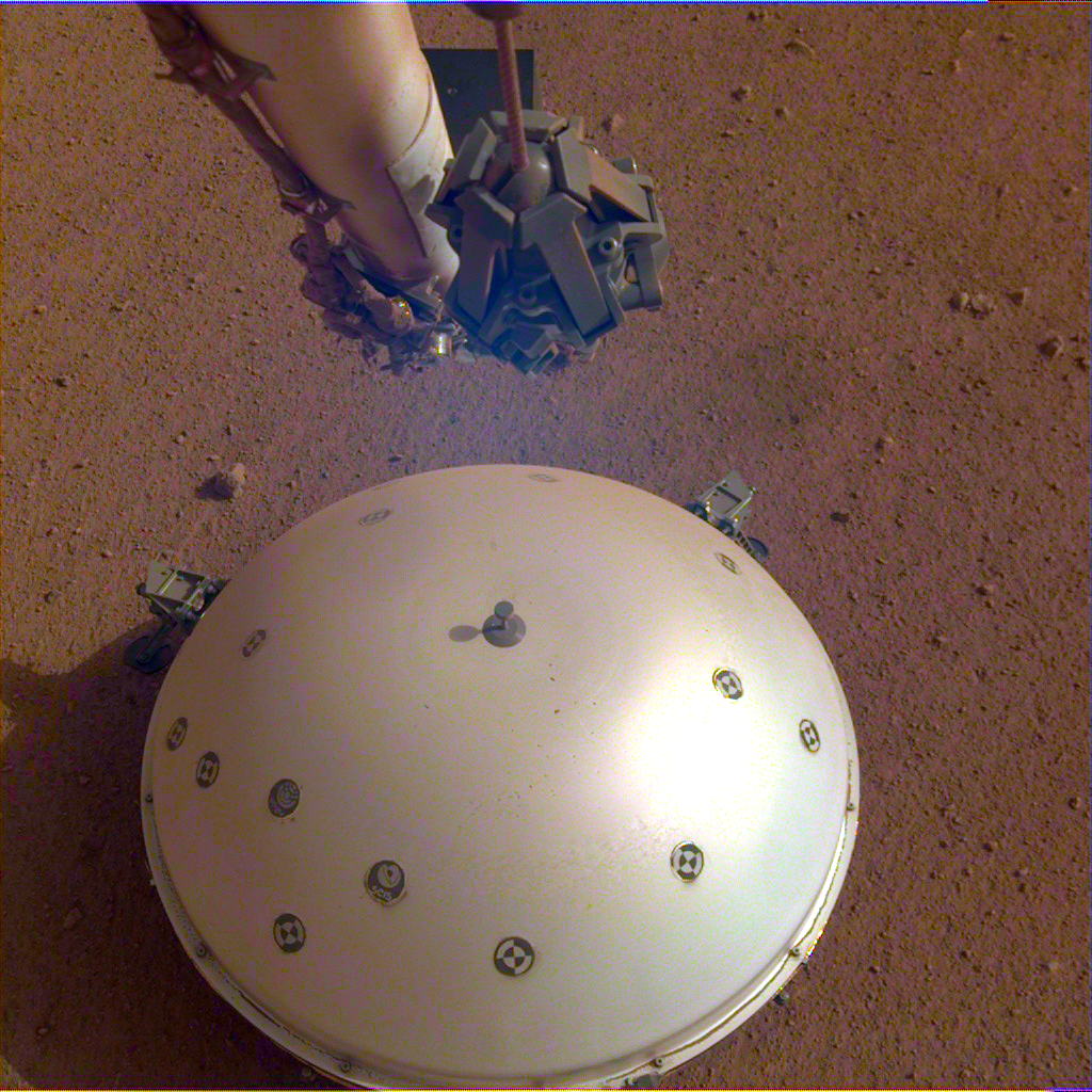 A robotic arm hovers above a small dome structure on the flat red surface of Mars.