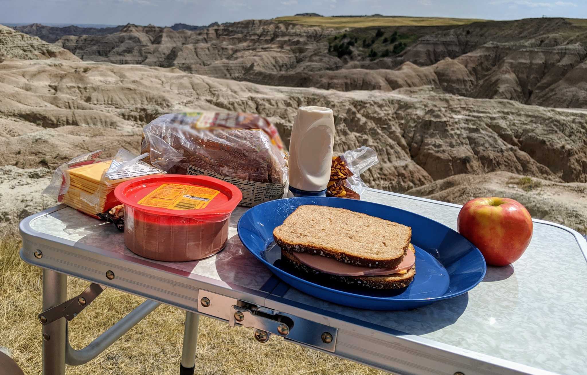 A table with the materials to make a sandwich, a sandwich, and an apple. The table is set out in front of a rocky landscape.