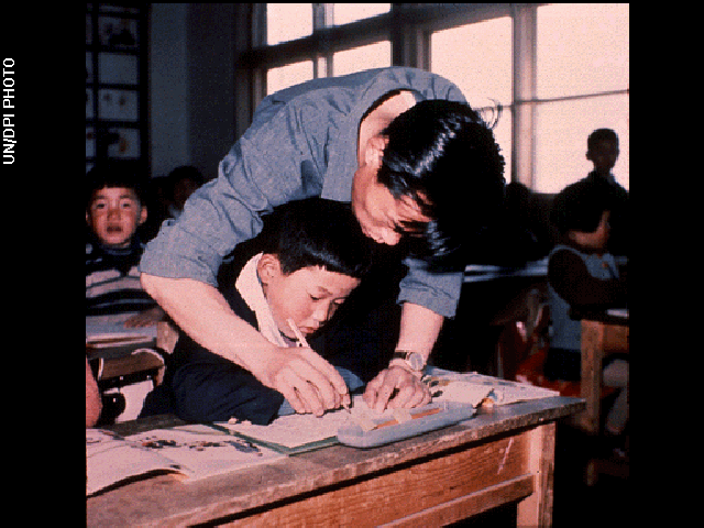 A teacher leans over his student's desk to help the child with writing on paper in a classroom