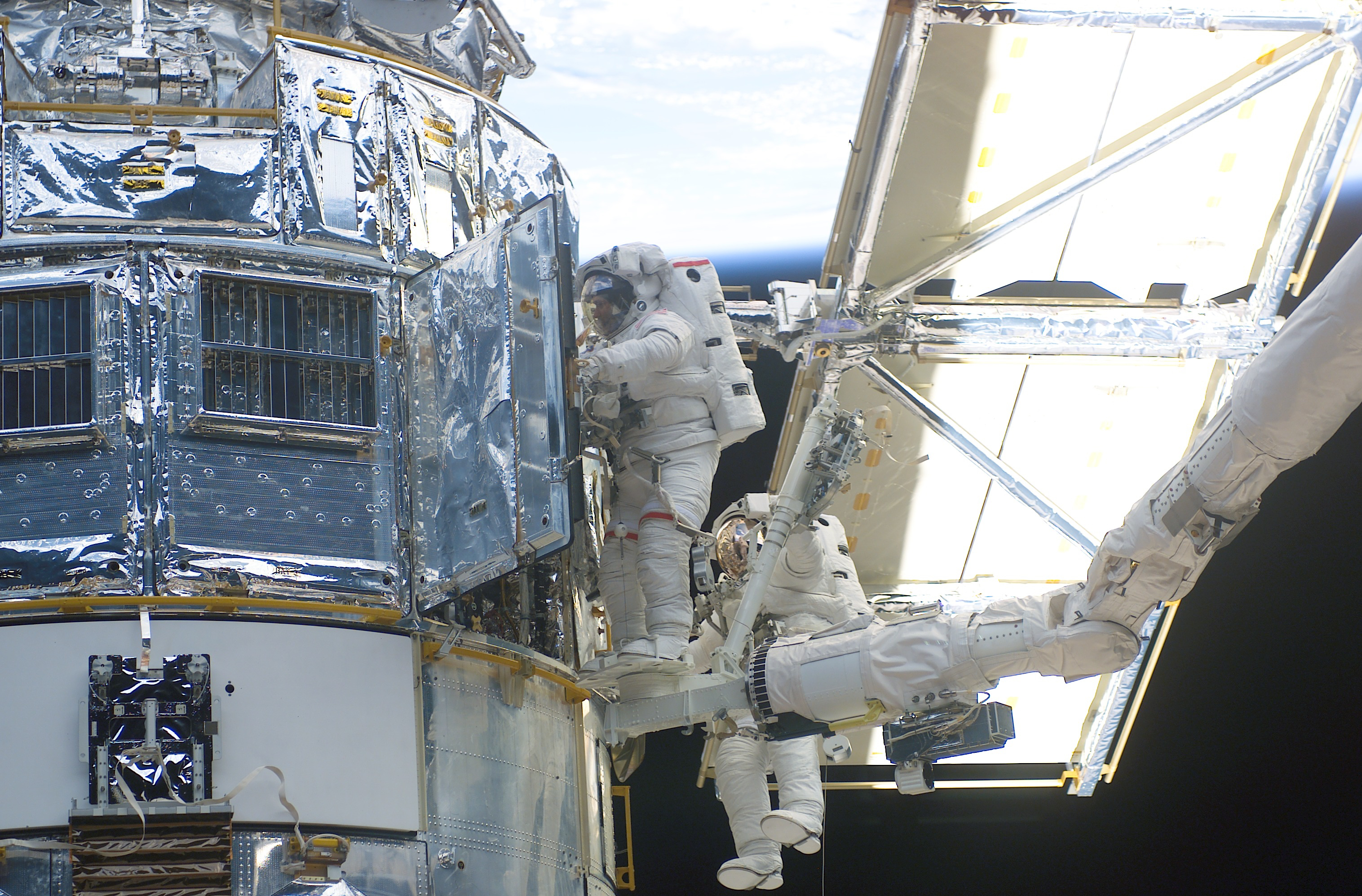 Two astronauts, one positioned on the space shuttle's robotic arm, work on the Hubble Space Telescope. One astronaut peers into an open door on the telescope. The telescope's solar array is clearly visible to the right.