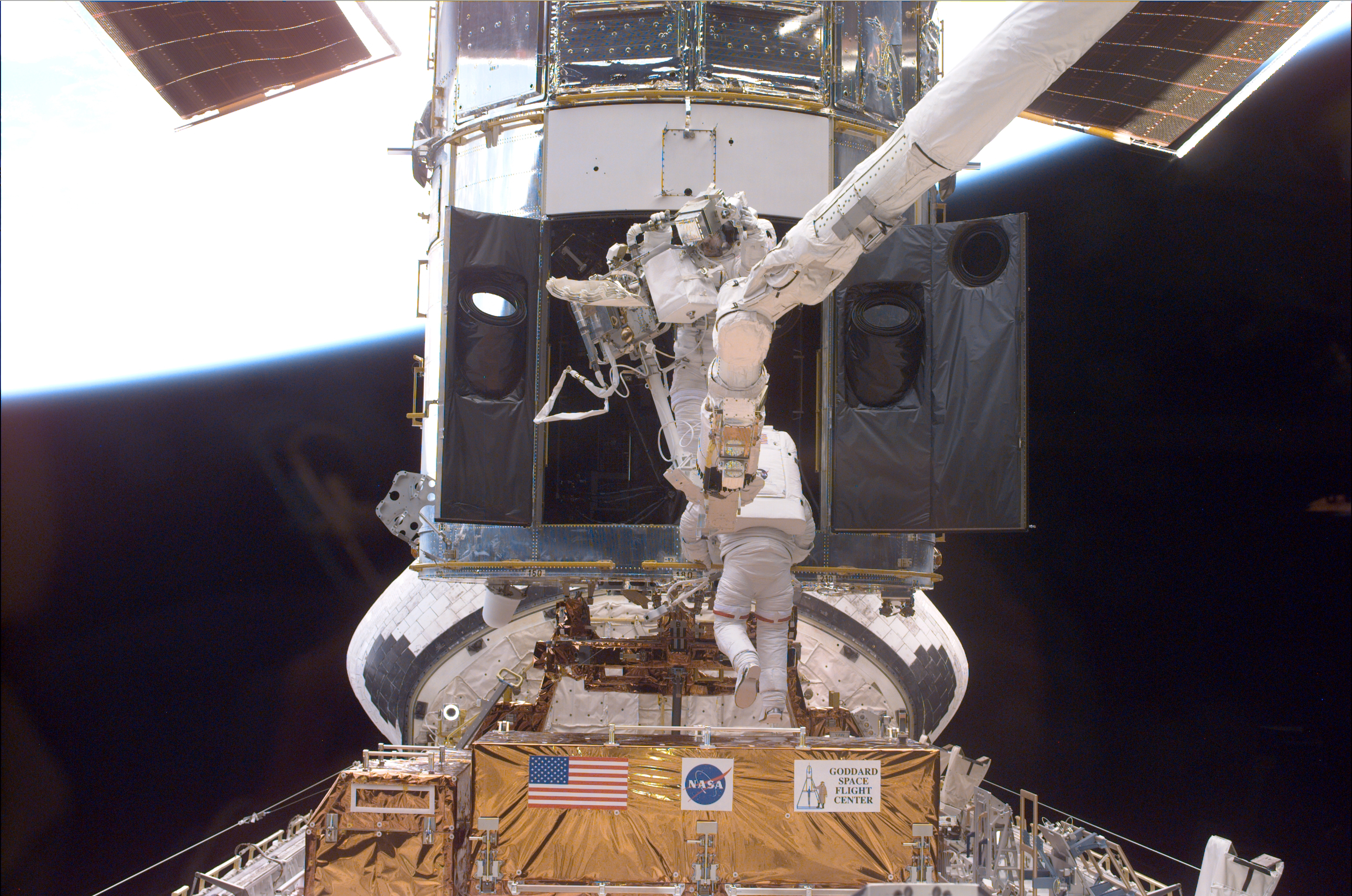 One astronaut is perched on the robotic arm and the other is gripping one of Hubble's handrails as they gaze into the wide open aft shroud doors.