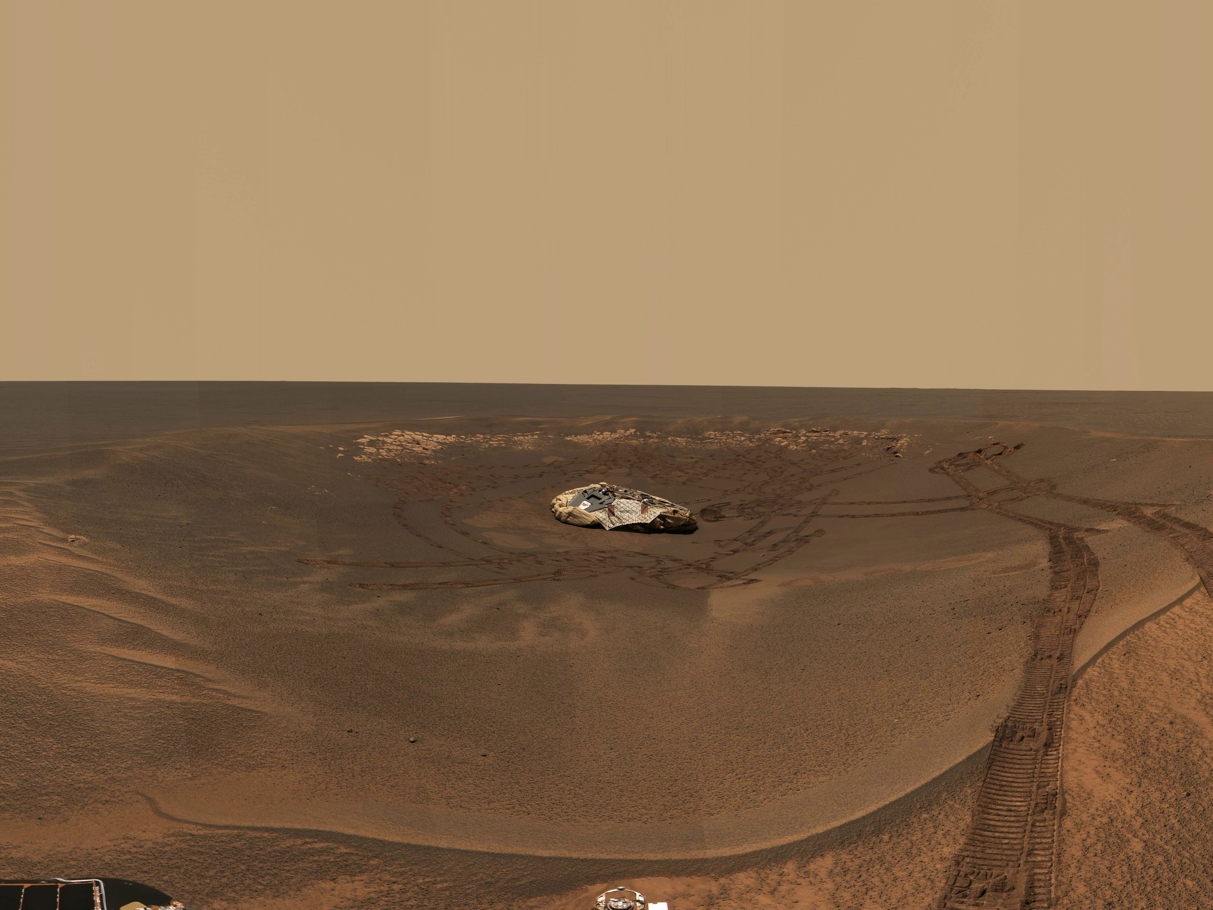 A landscape on Mars shows the horizon cutting across the middle of the photo, and a vast, empty desert of orange, brown, and rust stretching into the distance. The cloudless, hazy sky above is a pale tan color. In the foreground is a wide depression in the terrain, with a deflated, cream-colored bag lying in the middle, surrounded by wheel tracks, with one set running out of the crater toward the viewer and extending off the bottom of the image frame.