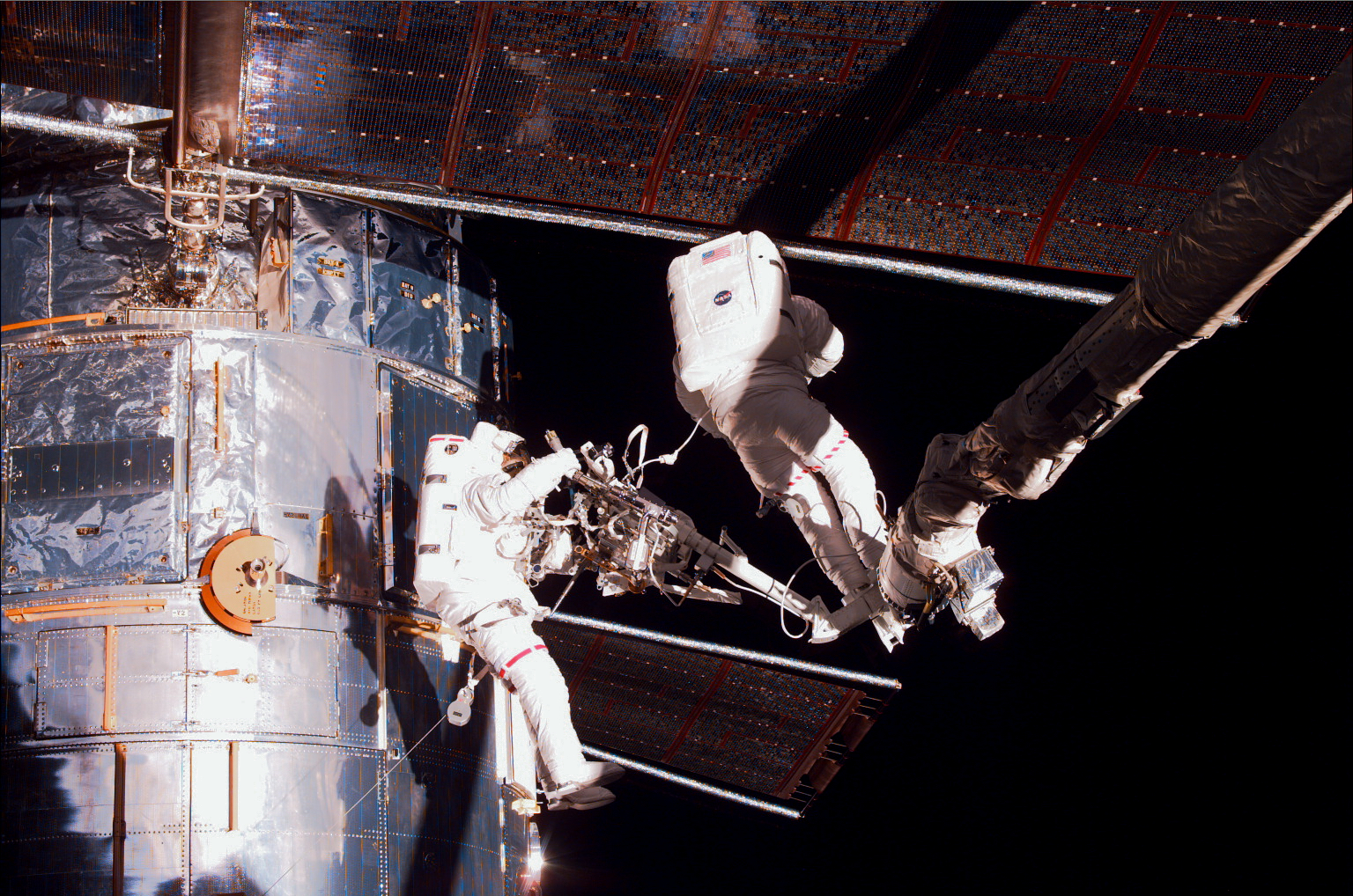 Two astronauts work on Hubble. The solar arrays are prominently visible. One astronaut is perched on the robotic arm.