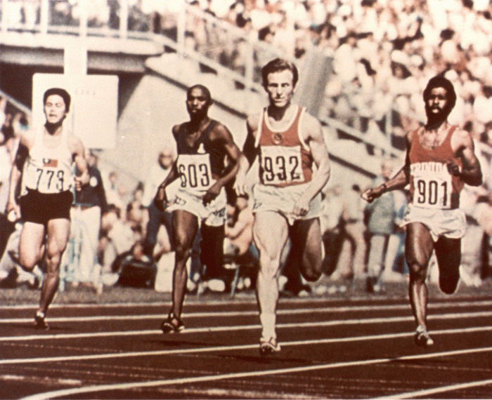 Four men in shorts running around a track in front of an audience at a competitive event