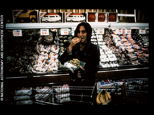 A woman stands with a cart at a supermarket, eating from a bag of green grapes she holds in her hand