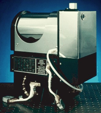 A boxy scientific instrument sits on an uncluttered surface. Its bottom half is dull black, metallic, and has a plaque with some writing affixed to its front. The upper half consists of two sections – on the right, a silver, metal box with crisp edges, about the same shape as a toilet tank. The left side is also metallic silver, with a curved top; the top has what looks like a semicircular door or indentation, resembling a swinging trash-can lid.