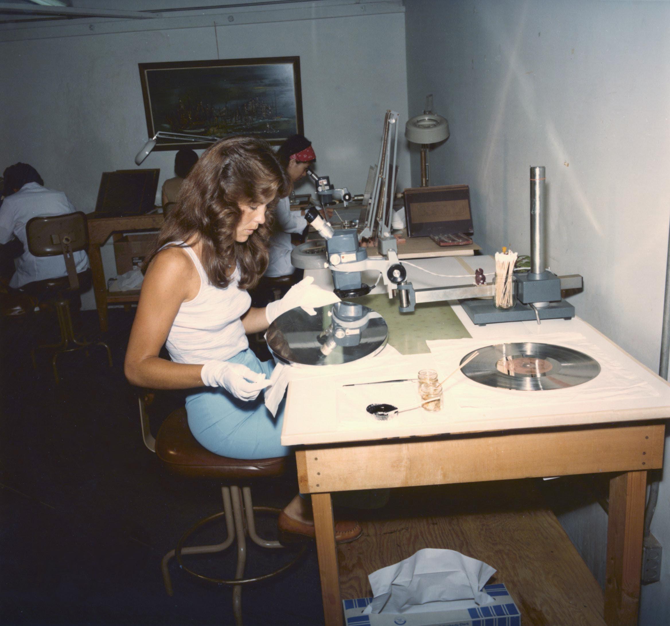 A woman wearing white gloves cleans a one of the discs in the Voyager golden record. A golden record is in the foreground.
