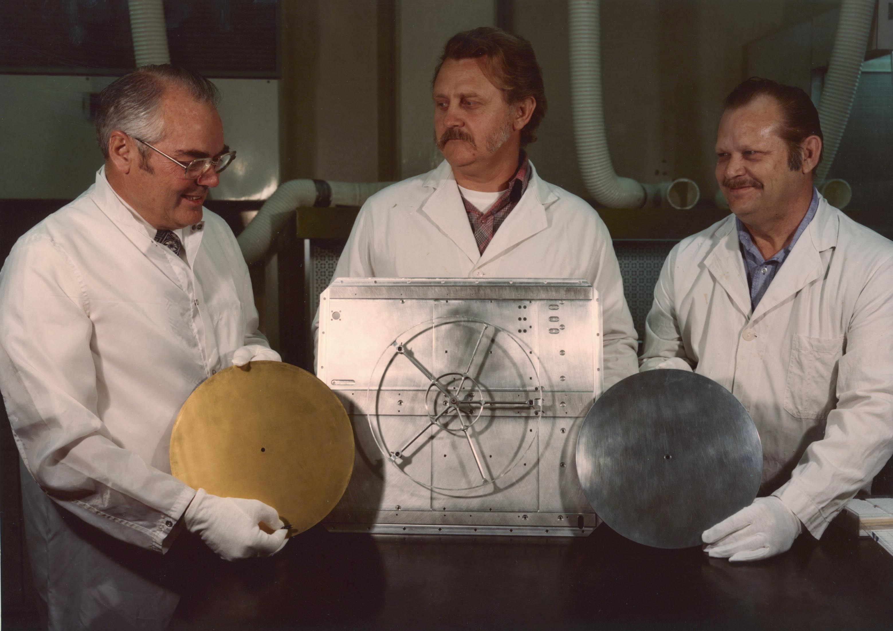 Three men display the unfinished components of the Golden Record, the cover, the spacecraft mounting bracket and the record.