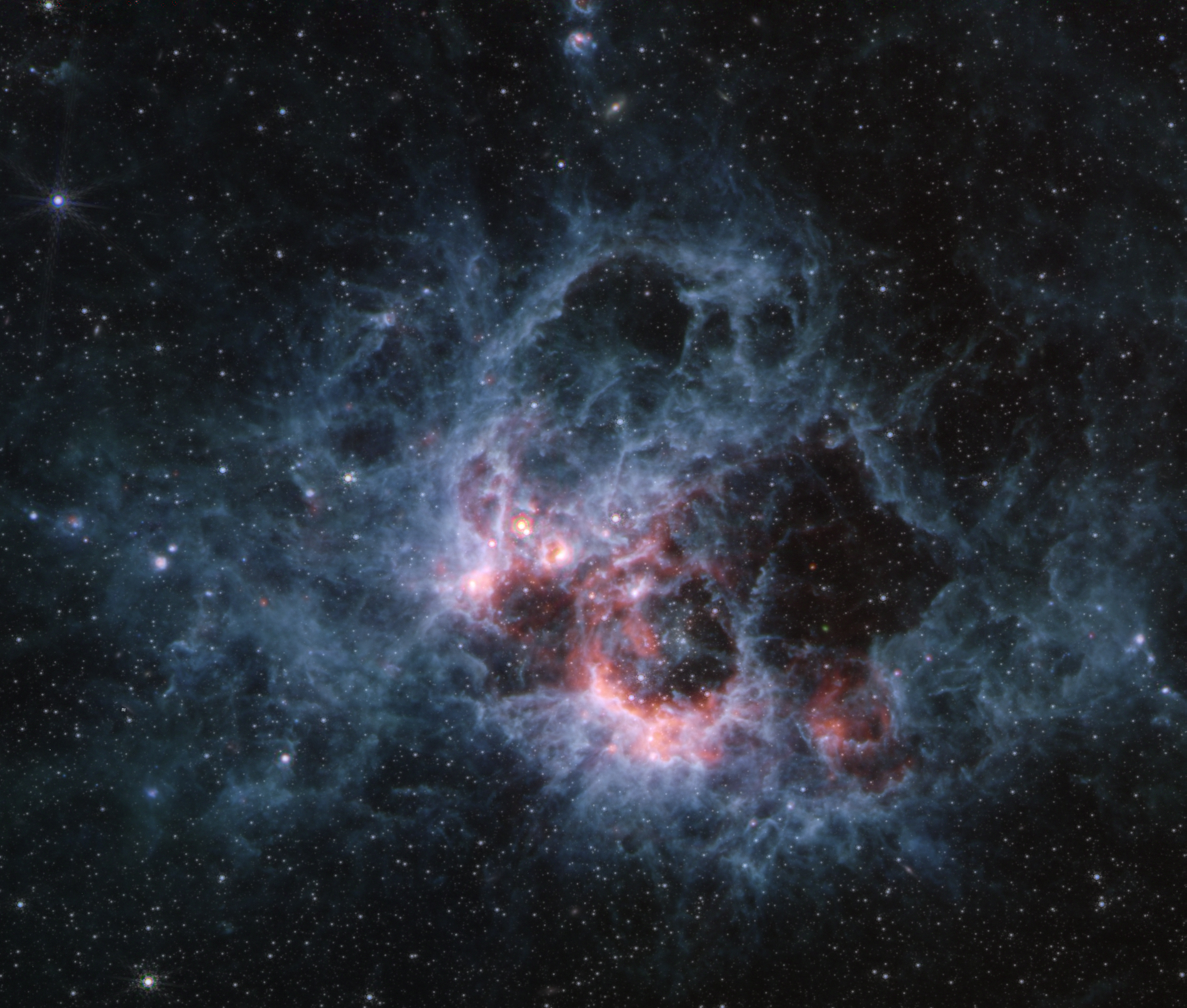 At the center of the image is a nebula on the black background of space. The nebula is comprised of wispy filaments of light blue clouds. At the center-right of the blue clouds is a large cavernous bubble. The bottom left edge of this cavernous bubble is filled with hues of pink and white gas. There are several other smaller cavernous bubbles at the top of the nebula, including two tiny cavities at the top center of the image. There are hundreds of dim stars that fill the surrounding area of the nebula.
