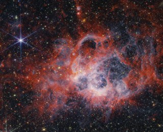 At the center of the image is a nebula on the black background of space. The nebula is comprised of clumpy, red, filamentary clouds. At the center-right of the red clouds is a large cavernous bubble, and at the center of the bubble there is an opaque blueish glow with speckles of stars. At the edges of the bubble, the dust is white. There are several other smaller cavernous bubbles at the top of the nebula, including two tiny cavities at the top center of the image. There are thousands of stars that fill the surrounding area outside the nebula, most of them are yellow or white. At 11 o’clock and 6 o’clock there are extremely bright stars with 8 diffraction spikes. There are also some smaller, <a href=