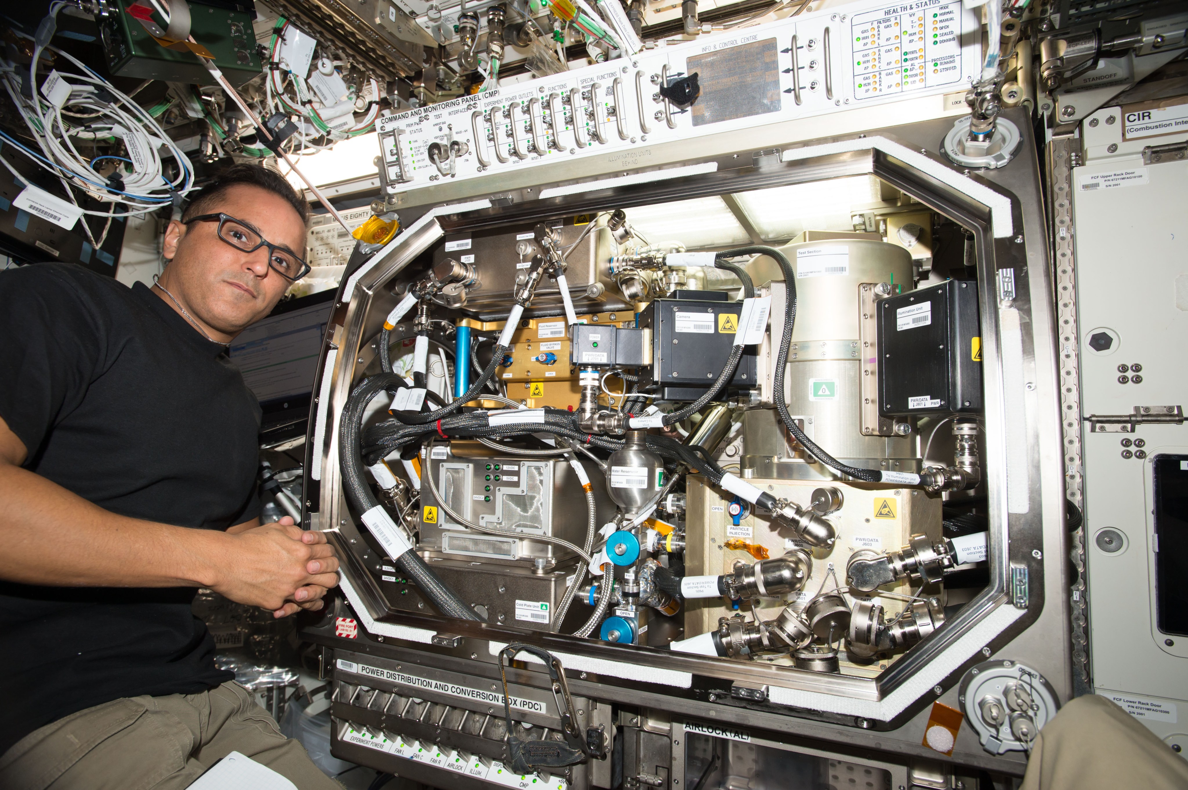 Astronaut Joseph Acaba wearing glasses and a black T-shirt is half standing, suspended in microgravity next to the ZBOT experiment in the Microgravity Science Glovebox (MSG) Unit aboard the station. The MSG is a rectangular compartment tightly fitted with various components including the test tank, enclosed in a cylindrical metallic vacuum jacket, sitting on top of a close Fluid Supply Unit (FSU) that is used for fluid thermal conditioning. The space in MSG is further crowded by a reservoir, various entangled hoses and wiring system, a camera and a small laser unit used for Particle Imaging Velocimetry (PIV) diagnostics that measures and visualizes fluid motion in the tank.