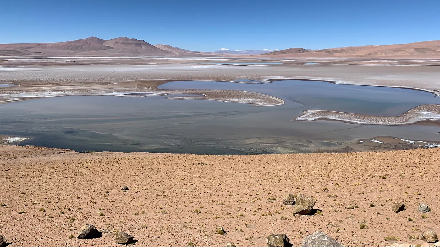 一幅风景画展现在一个几乎平坦的表面上，表面上有许多液体池。在中间，远处的山脉穿过图像的顶部，其间点缀着日光蓝色的天空。