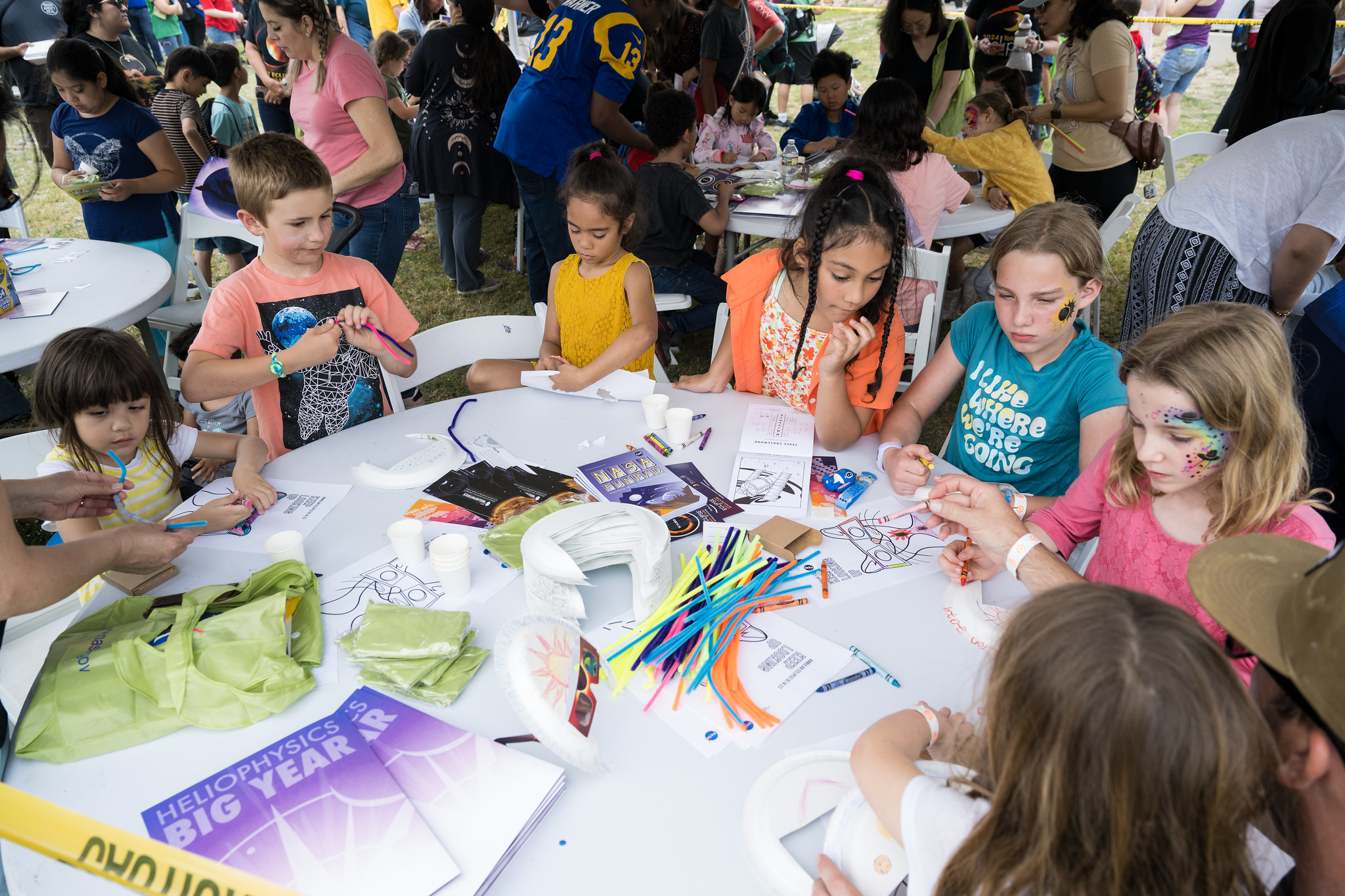 Several children stand around a large white circular table. Art supplies are scattered across the table, including pipe cleaners, paper plates, coloring sheets, posters, and crayons.
