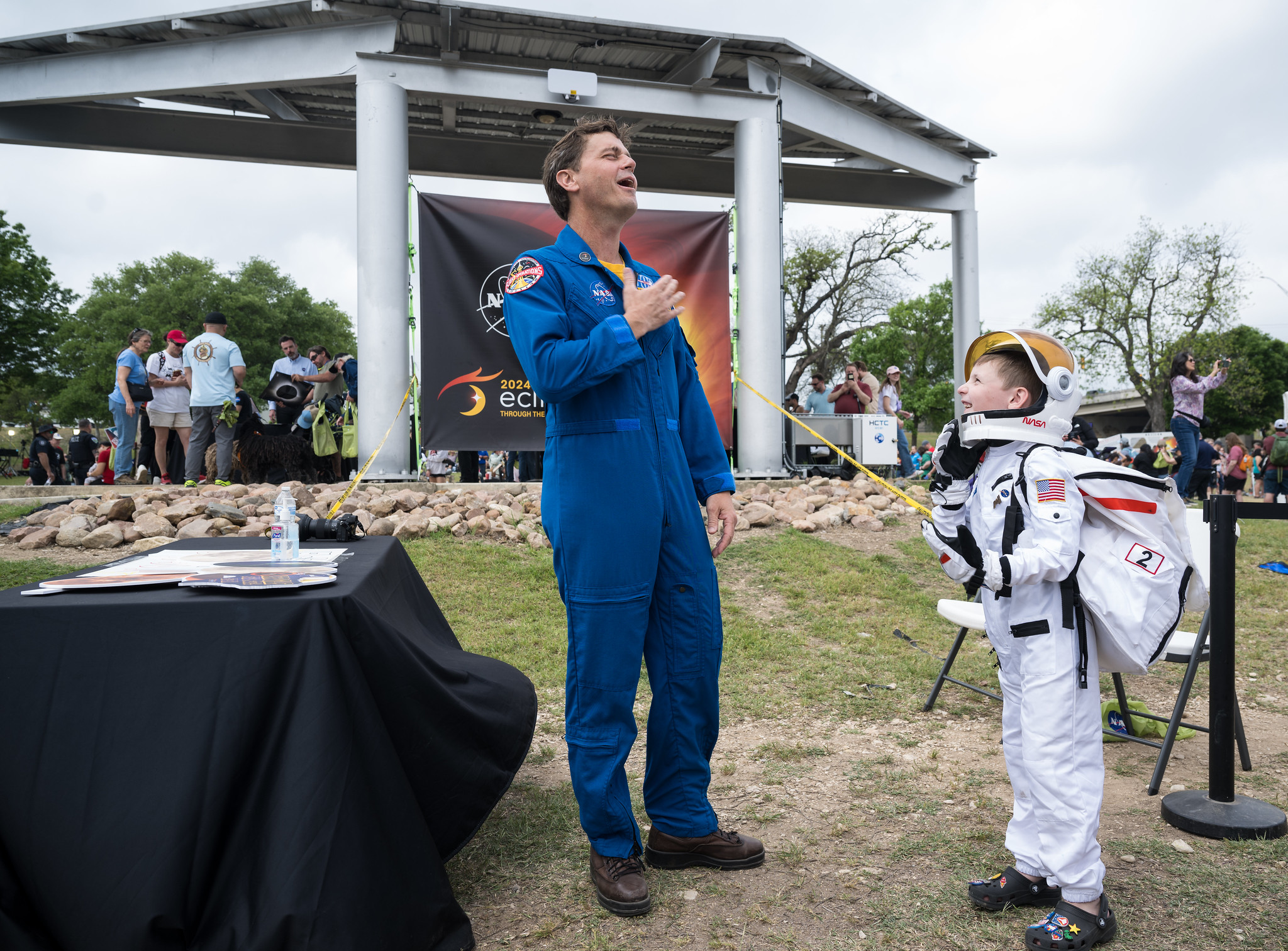 A man in a blue astronaut jumpsuit stands on grass next to a young child wearing an astronaut costume. They are outdoors, near a table with eclipse-themed photo props. Behind them, people stand under a white pavilion. There is a large banner on the pavilion that says 