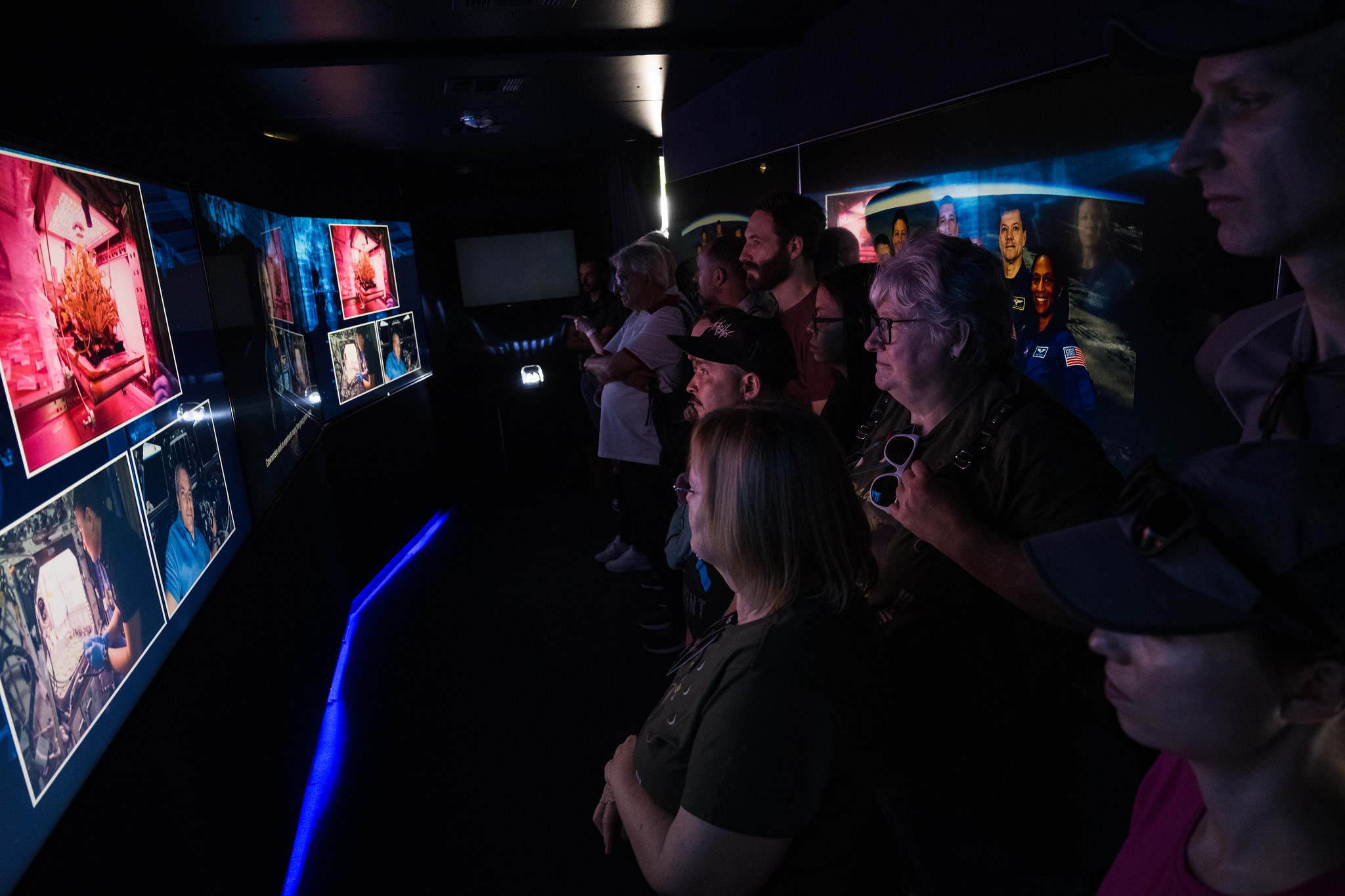 In a darkened room, several people stand in front of a collection of screens. The screens show images of the inside of the International Space Station. The light from the screens is reflecting off the visitors' faces.