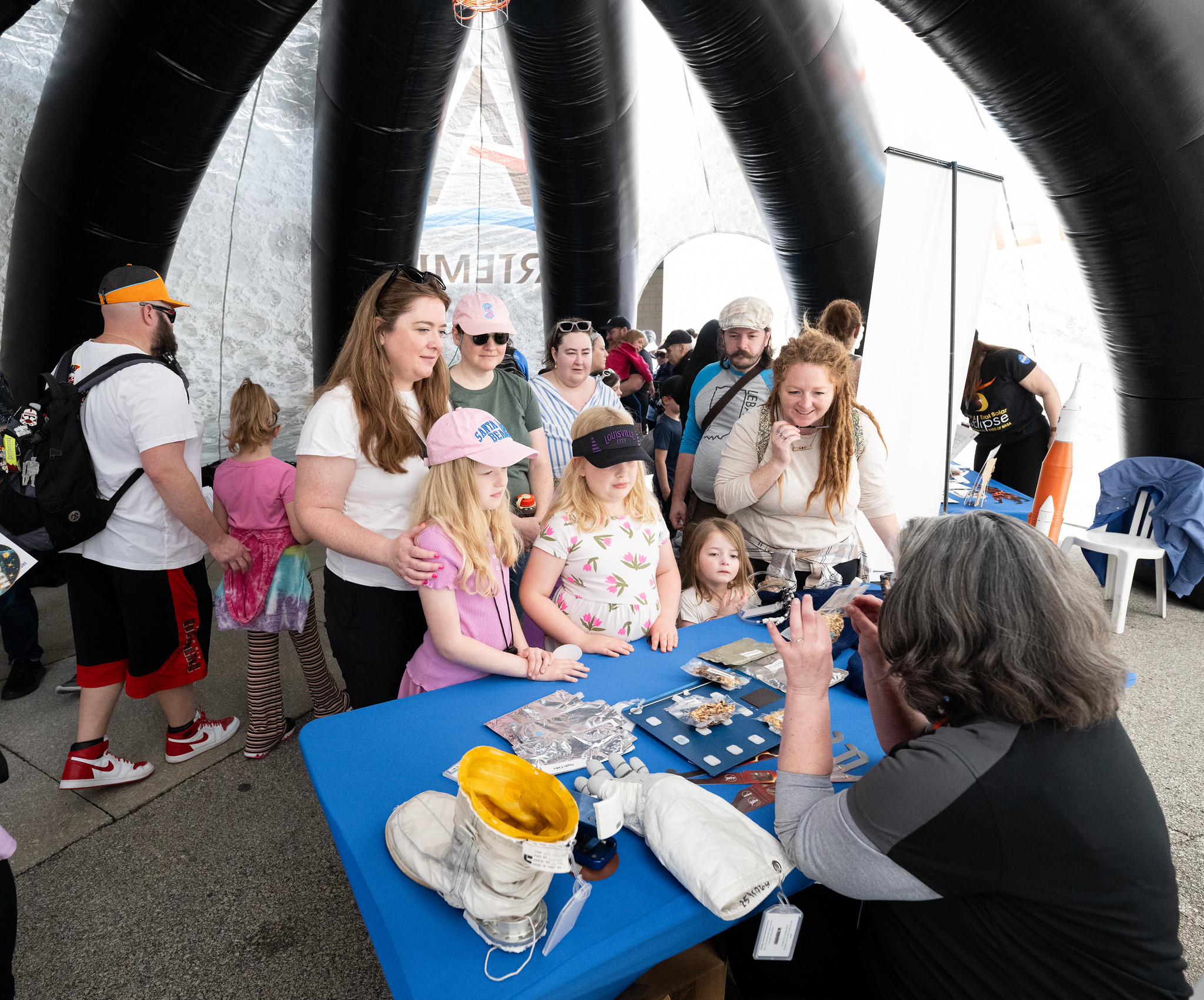 A group of children and adults stand in front of a table. On the other side of the table a person is holding a small clear bag with small brown, indistinguishable objects in side. On the table are an astronauts boot, gloves, eclipse glasses, and other indistinguishable items.