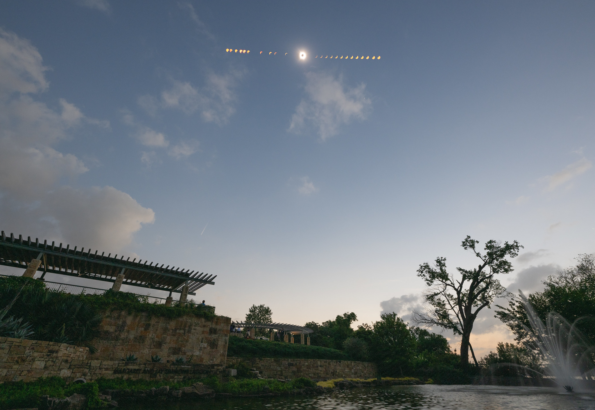 Above a garden, pergola, pond, and fountain, the stages of the eclipse are seen in the sky. From left to right, the Sun changes from partially covered, to a crescent, to a total solar eclipse, then back to a crescent and partially covered again.