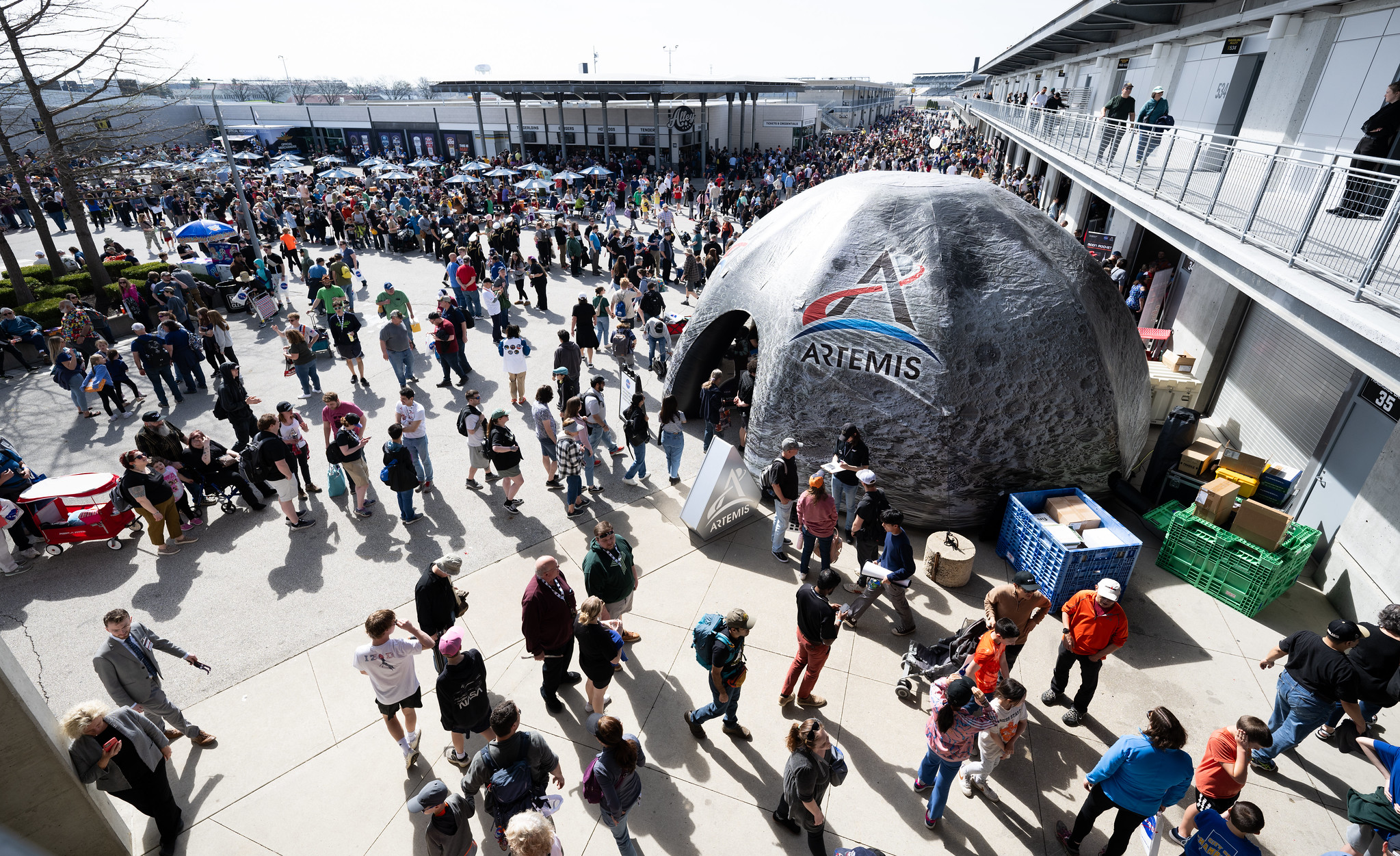A view from above of a gray, large dome tent with images of the Moon. The tent has the Artemis insignia on it. Crowds of many people stand around the outside of the tent.