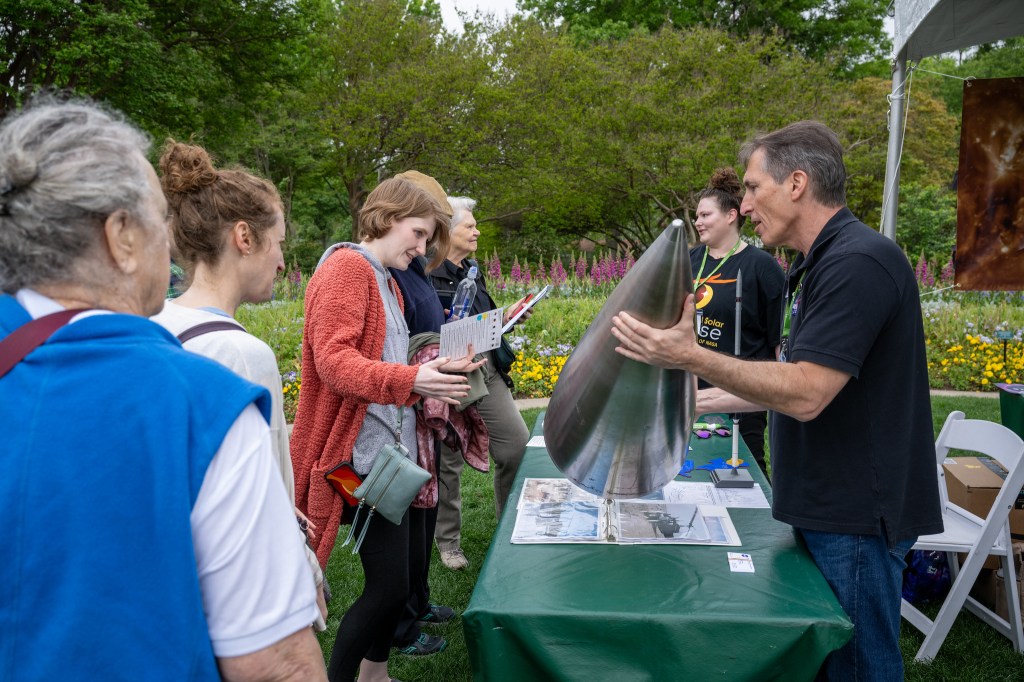 A man stands next to a woman at a table with a green table cloth. He is using both hands to hold a large, silver metal cone. He is talking. Several people stand on the other side of the table, looking at the cone.