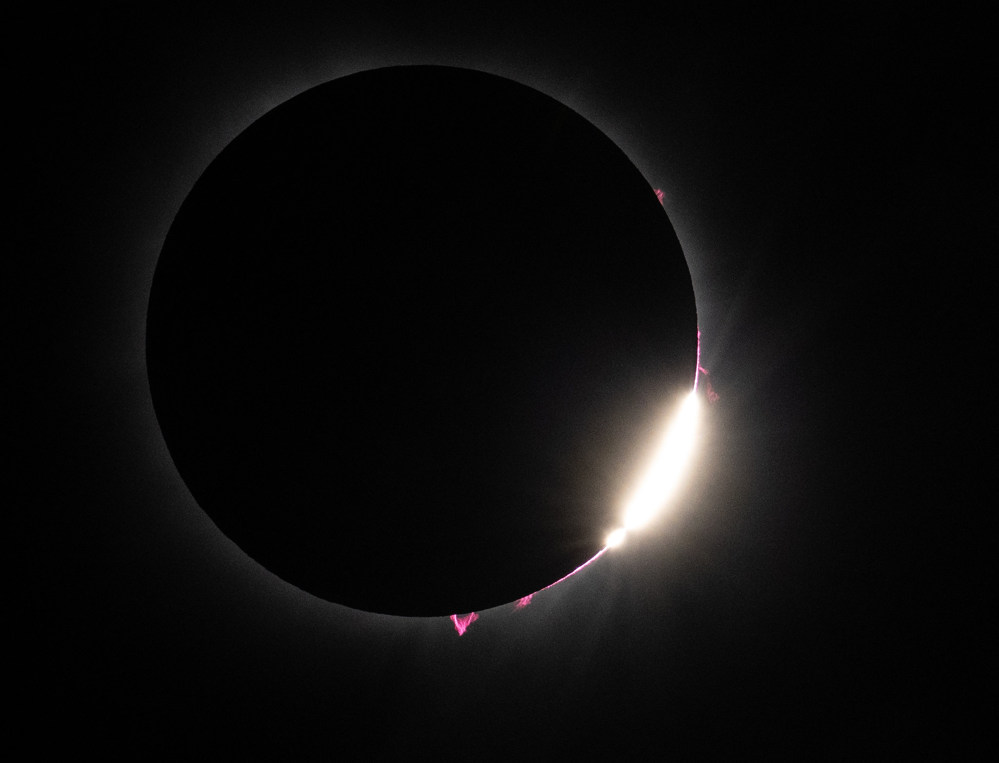 The total solar eclipse. A large black circle against a black background. A faint white glow is seen around the rim of the black circle. On the bottom right, a bright burst of light peeks out. Toward the center of the bottom of the circle, a bright pink, slightly transparent bit of solar matter flows against the black background. A few other spots of bright pink material can be seen on the right area of the circle.