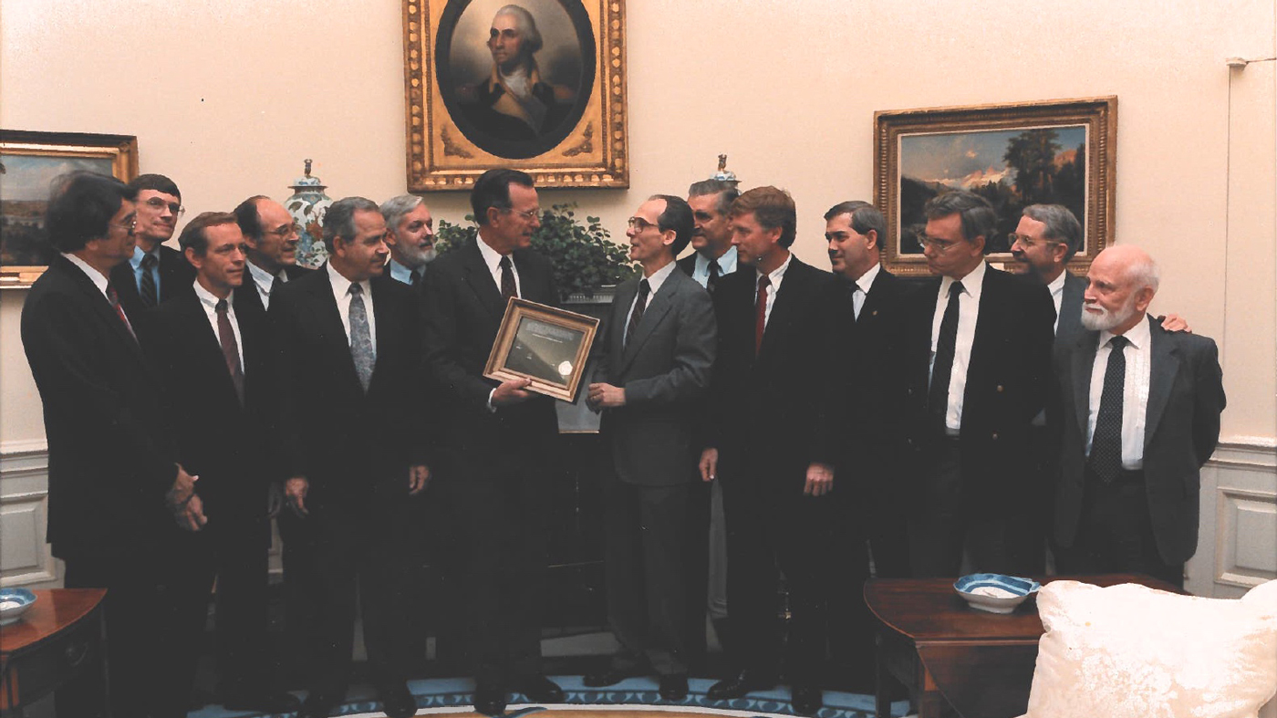 A group of men stand around President Bush as he receives a framed photo from a scientist.