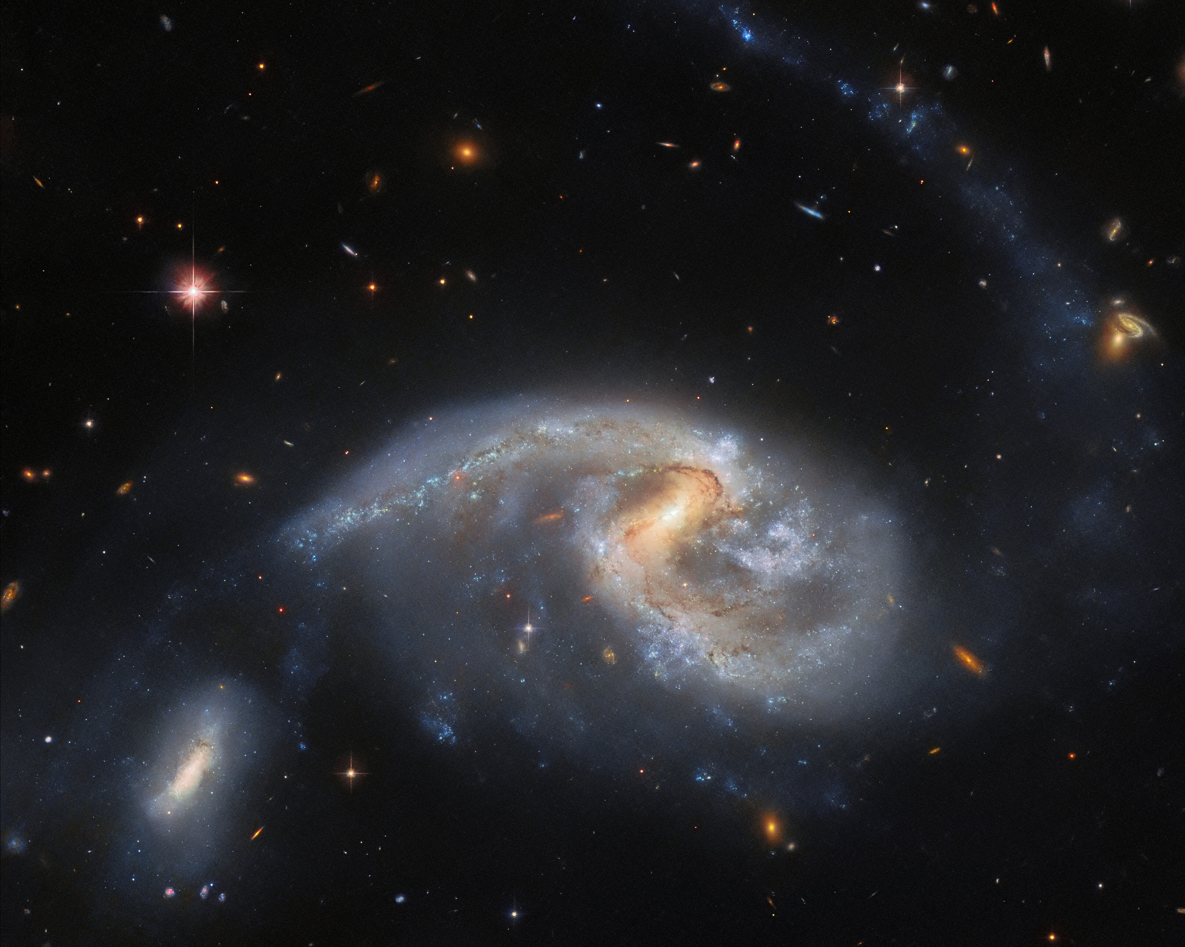 Una gran galaxia espiral con una galaxia vecina más pequeña. La galaxia espiral es ancha y distorsionada, con polvo de colores. Su compañera se encuentra cerca, al final de un brazo en espiral, en la parte inferior izquierda. Una larga y tenue cola de estrellas se extiende desde el lado derecho de la galaxia espiral hasta la parte superior de la imagen. Varias galaxias pequeñas y distantes son visibles en el fondo, junto con una estrella brillante en primer plano.